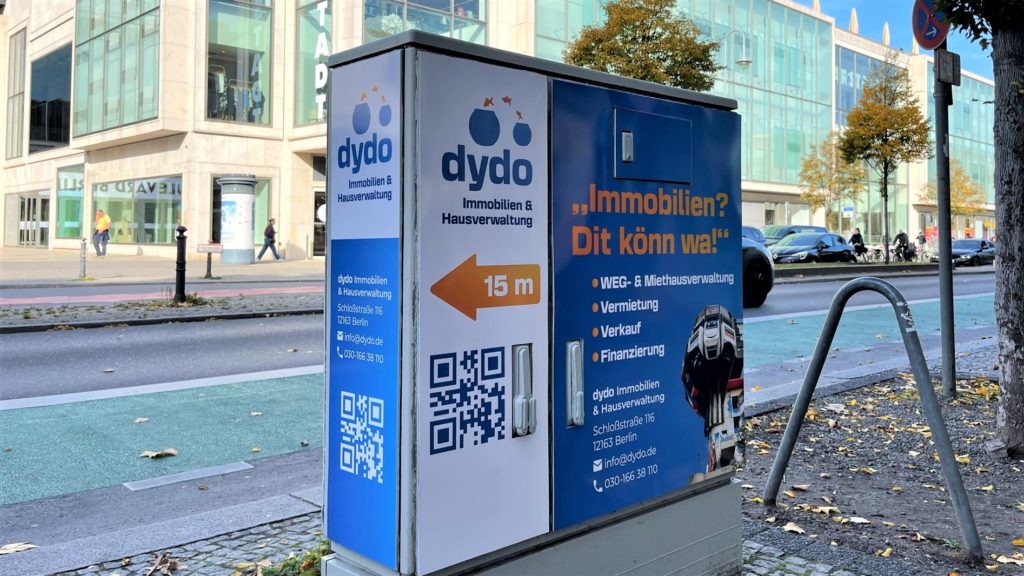 Nahaufnahme einer Stromkastenwerbung für Dydo. Der Stromkasten zeigt eine Wegbeschreibung zum Ladengeschäft.