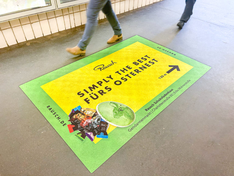 Bodenfolie in einem Zugang zur U-Bahn. Die Außenwerbung ist grün-gelb und wirbt für "Rausch-Schokoladen".