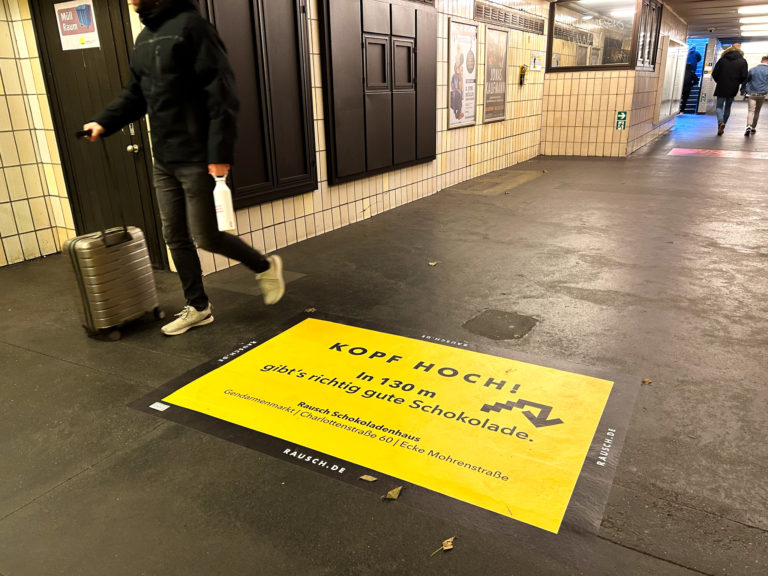 Fußbodenwerbung in einem U-Bahnhof mit Wegbeschreibung in einem knalligen Gelbton.