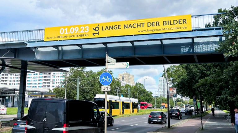 Ein gelbes Brückenbanner hängt von einer Brücke. Im Hintergrund sind Autos und eine Tram.