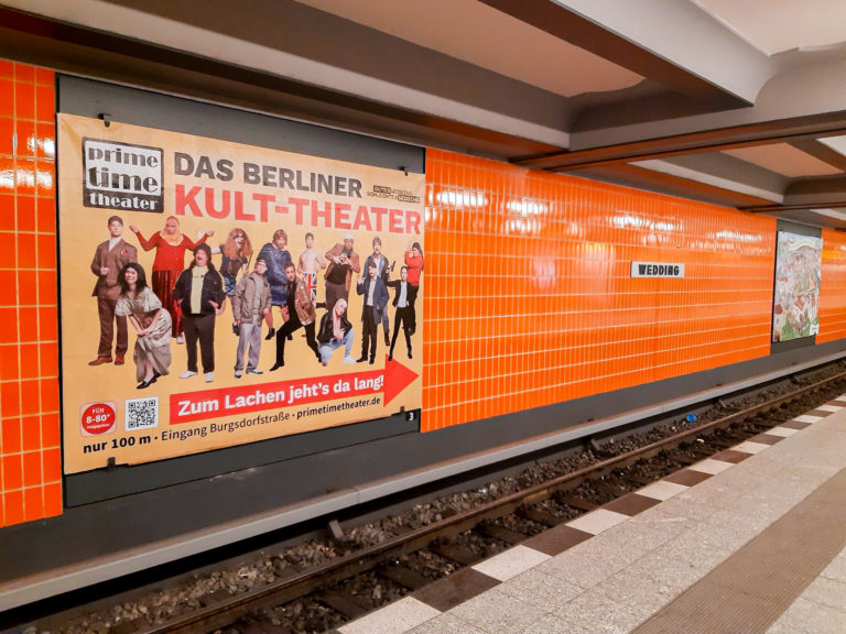 Ein U-Bahnhof in orange mit Großflächen-Werbung für ein Berliner Theater.
