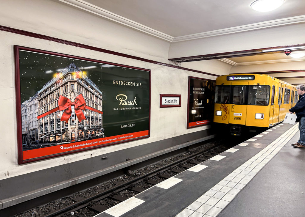 Auf einem Bahnhof fährt gerade die gelbe U-Bahn ein. Im Vordergrund ist eine weihnachtliche Werbung für Schokoladen-Rausch.