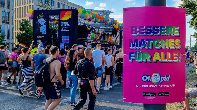 Hohlkammerplakat als Außenwerbung auf einem Straßenfest in Berlin. Das pinke Plakat hängt an einem Masten. Im Hintergrund sind viele Menschen unterwegs.