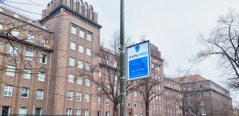 Ein blau-weißes Mastenschild hängt an einer Laterne. Im Hintergrund ist ein Backsteingebäude.