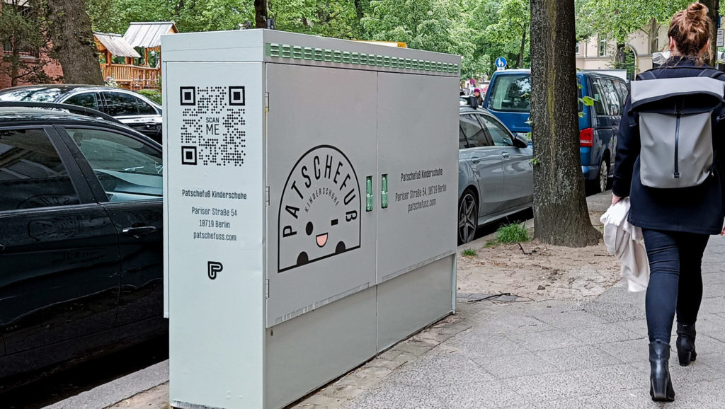 Ein großer grau-grüner Stromkasten wirbt für ein Schuhgeschäft in der Nähe. Auf dem Gehweg läuft gerade eine Passantin, mit dem Rücken zur Kamera, vorbei.