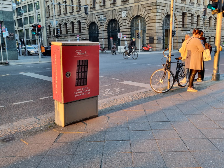 Auf einer Straße steht ein beklebter Stromkasten, in rot. An der Ampel stehen Passanten.