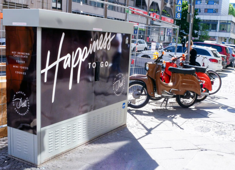 Auf einem brauen Stromkasten steht "Happiness". Die Stromkastenwerbung ist für Schokoladen-Rausch. Ein Mopped steht rechts vom Kasten.