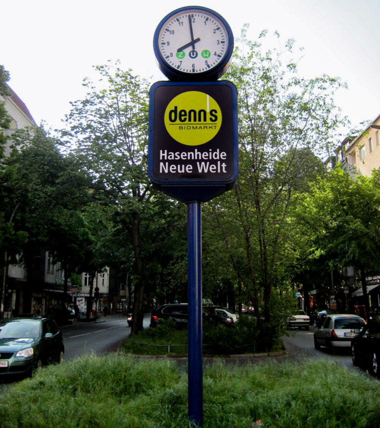 Uhrenwerbung mit einem Logo von Denns. Die Werbeuhr steht auf einem Grünstreifen.