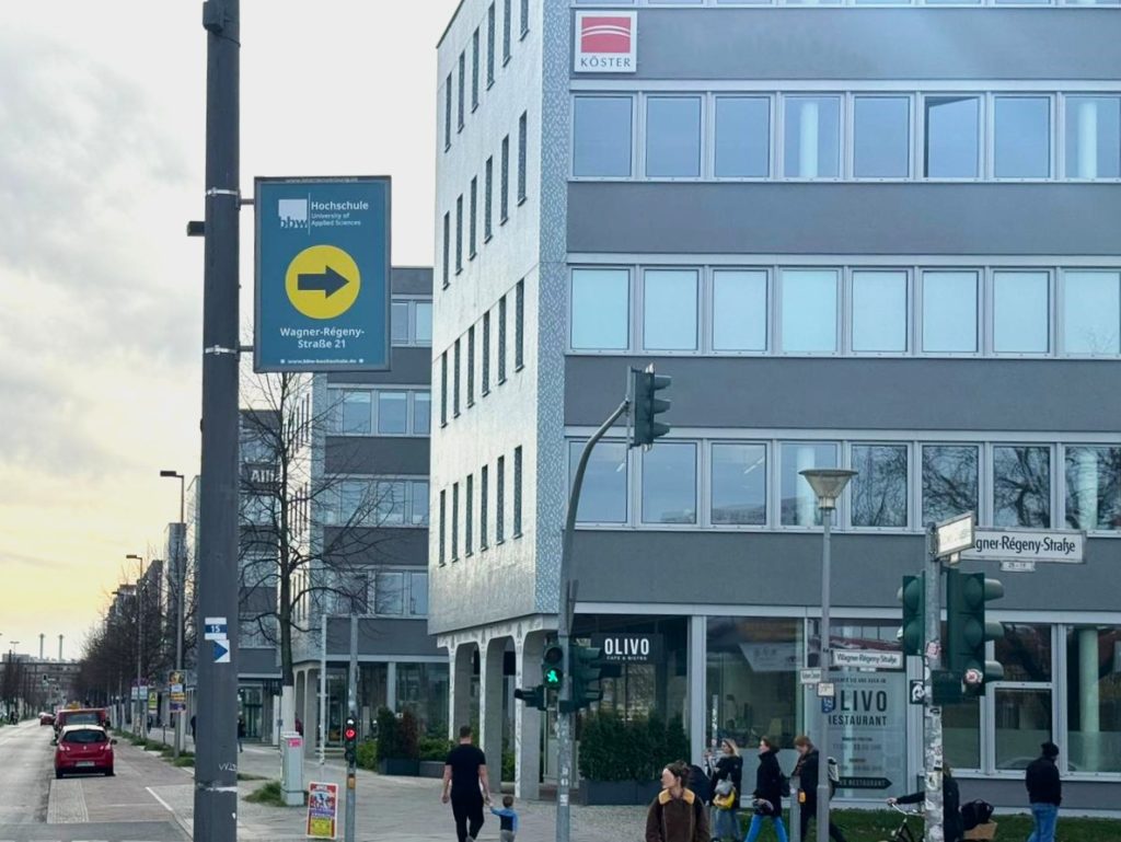 In Adlershof steht an der Ecke Rudower Chaussee Ecke Wagner-Régeny-Straße ein Lichtmast, an dem ein Mastenschild der BBW Hochschule hängt. Das Schild weist den Weg zum Hochschulstandort mit einem großen Pfeil. Im Hintergrund sieht man eine breite Straße und ein Bürogebäude. Im Vordergrund überqueren Menschen die Ampel.