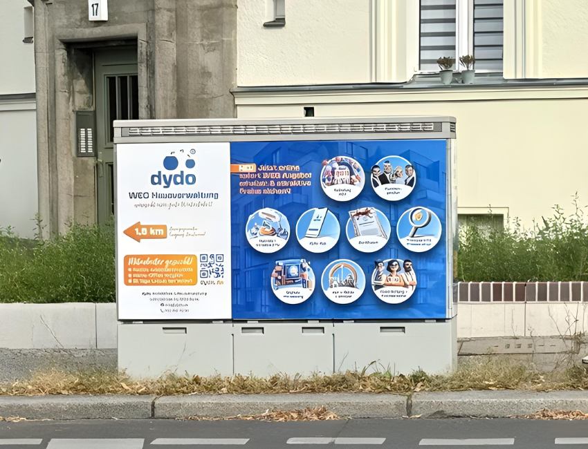 Seitens der Filanderstraße steht ein Telekomkasten der die Außenwerbung der Dydo zeigt. Auf dem Motiv sieht man die einzelnen Dienstleistunden die sie anbieten, das Logo und einen Standorthinweis. Im Hintergrund des Werbeträgers ist der Gehweg, sowie ein Mehrfamilienhaus.