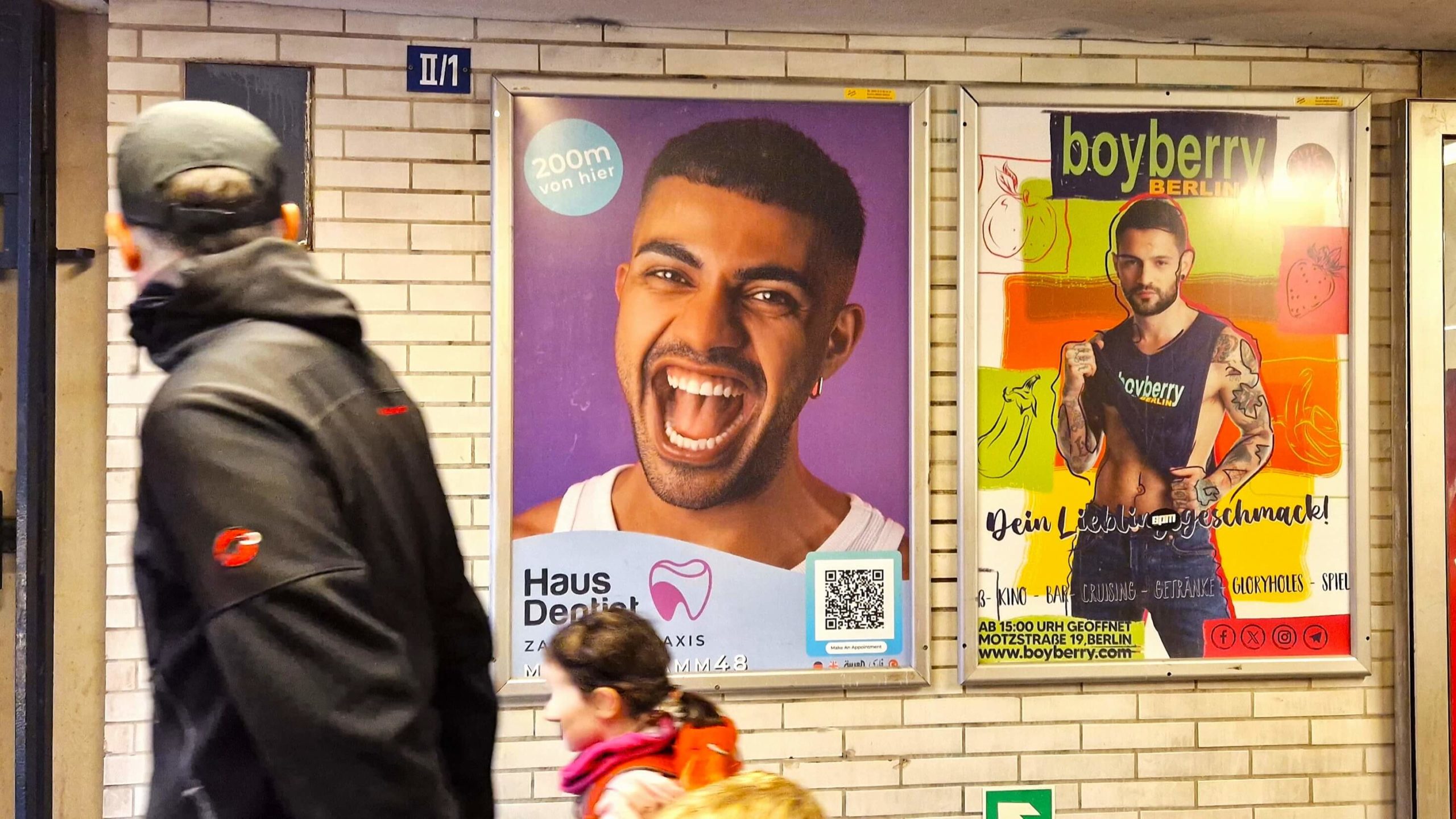 An einer Wand am U-Bahnhof Mehringdamm hängt ein A0 Plakat der Zahnarztpraxis Haus Dentist. Auf der Sonderfläche ist ein fröhlich lächelnder Mann zusehen. Die Außenwerbung verweißt auf den Standort der Zahnarztpraxis. Neben dem Plakat ist eine weitere Fläche zu sehen. Ein Mann mit Kind läuft an der Plakatwerbung vorbei.