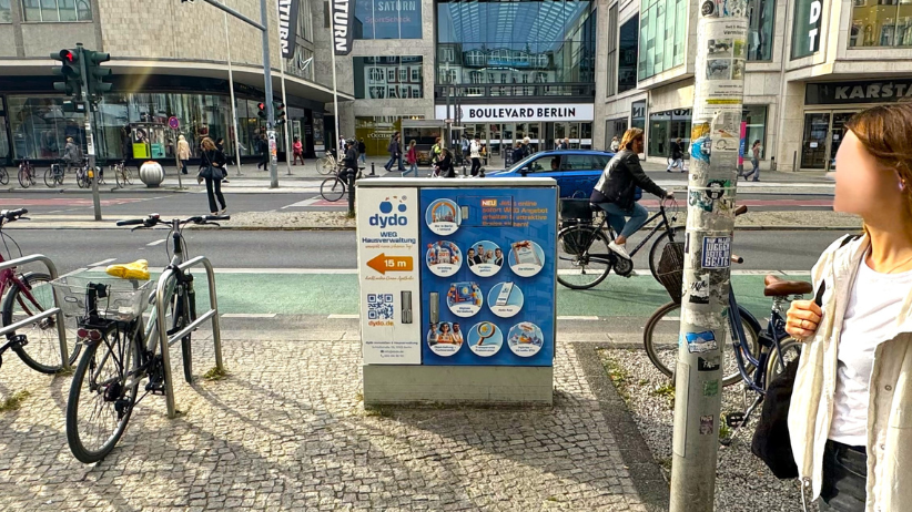 An der Schloßstraße in Steglitz steht ein Ampelkasten mit der Werbung von Dydo. Die Außenwerbung zeigt direkt zu ihrem Büro und weist die Leistungen aus. Hinter dem Werbeträger sieht man die Hauptstraße und den Boulevard Berlin. Um die lokale Werbung sieht man Fußgänger, Autofahrer und Fahrräder.