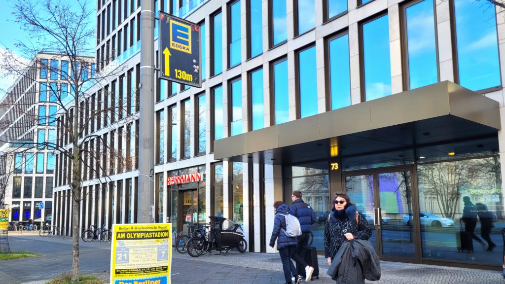 Edeka wirbt in der Invalidenstraße 73 auf einem Mastenschild. Die Laternenwerbung weist die Kunden zum Einzelhandel. Im Hintergrund sind Gebäude und Passanten zu sehen.