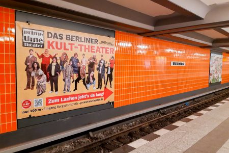 Ein U-Bahnhof in orange mit Großflächen-Werbung für ein Berliner Theater.