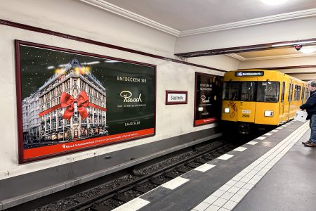 Auf einem Bahnhof fährt gerade die gelbe U-Bahn ein. Im Vordergrund ist eine weihnachtliche Werbung für Schokoladen-Rausch.