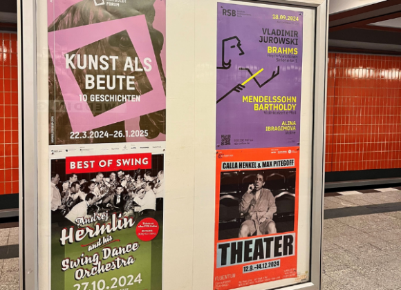 Eine Plakatvitrine, die vier Plakate zeigt, steht an einem Bahnhof. Das rote Plakat von Fluentum befindet sich unten rechts in der Vitrine.